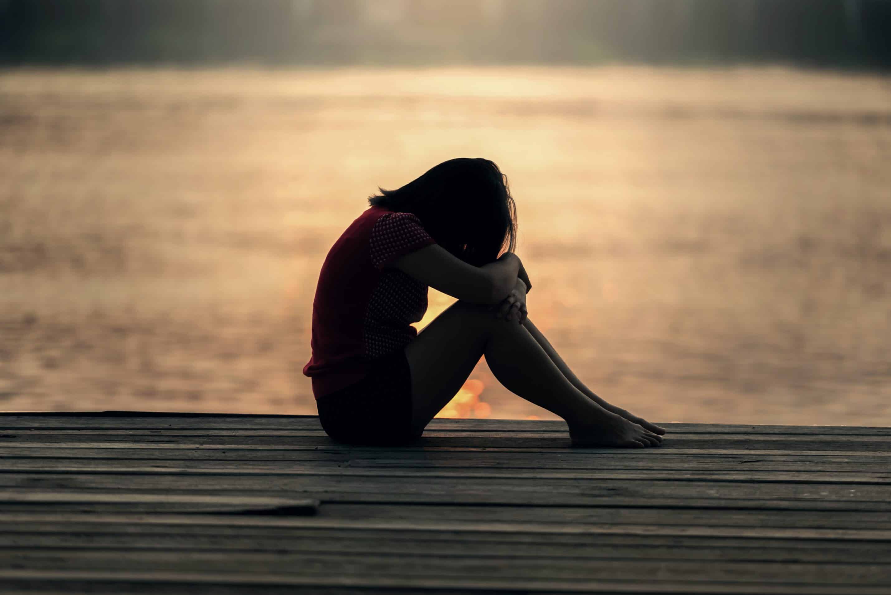 Woman with head on knees in front of water and sunset.